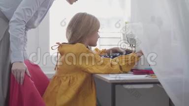 小女孩学着如何缝制坐在<strong>靠窗</strong>的<strong>桌子</strong>旁的衣服，而她的红头发的母亲在帮助和帮助